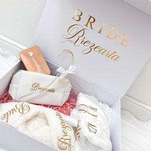 Może przedstawiać: Białe pudełko prezentowe ze złotym napisem „BRIDE Prezearta”. W środku biały szlafrok, kosmetyczka i kompaktowe lusterko. Szlafrok i torba mają złote napisy. Pudełko wypełnione jest różową pociętą bibułą.
