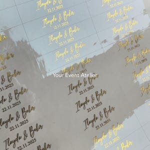 Puede incluir: Pegatinas doradas con los nombres "Ilayda & Bekir" y la fecha "22.11.2025" impresos en una elegante escritura. Las pegatinas están dispuestas en filas y columnas sobre un fondo blanco. El texto "Your Event Atelier" también es visible.