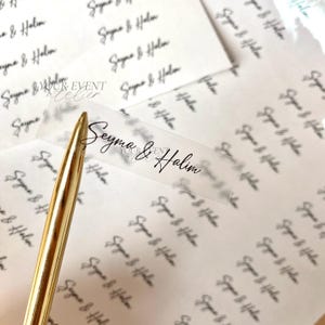 Könnte beinhalten: Nahaufnahme von transparenten Etiketten mit den Namen "Seyma & Halim" in schwarzer Schrift. Ein goldener Stift ist im Vordergrund. Der Hintergrund ist mit demselben Namen gefüllt.