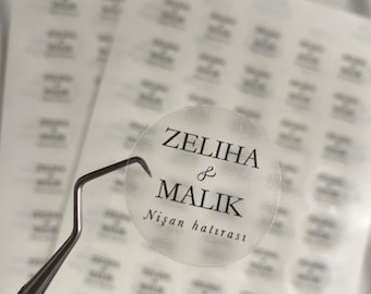 Pode incluir: Um adesivo redondo transparente com texto preto que diz "Zeliha & Malik Nişan hatırası". O adesivo está em uma folha de adesivos transparentes.