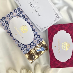 Può includere: Una scatola regalo piena di cioccolatini, decorata con un motivo blu e bianco. La scatola ha una carta allegata con il testo "Eid Mubarak" in lettere dorate. Sono visibili anche due carte aggiuntive con il testo "Eid Mubarak", una bianca con lettere nere e una rossa con lettere dorate.