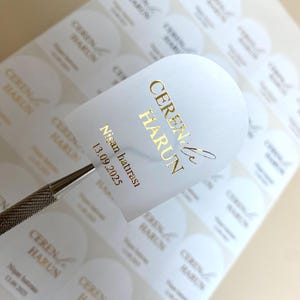 Könnte beinhalten: Weiße, bogenförmige Aufkleber mit goldfarbenem Text. Der Text lautet "CEREN & HARUN" und "Nişan hatırası 13.09.2025". Ein Metallwerkzeug hält einen Aufkleber, viele weitere befinden sich im Hintergrund.