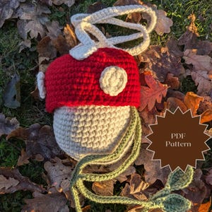 May include: A crocheted mushroom-shaped bag with a red top, white spots, and a long green strap. The bag is lying on a bed of brown leaves. PDF Pattern.