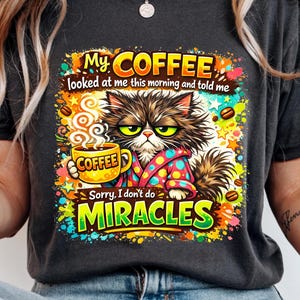 Puede incluir: Camiseta gris oscuro con un gráfico colorido de un gato gruñón sosteniendo una taza de café. El texto dice: "My COFFEE looked at me this morning and told me Sorry, I don't do MIRACLES."