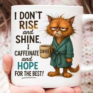 May include: White ceramic mug with a humorous design. The mug features a grumpy cartoon cat in a teal robe holding a coffee cup. Text on the mug reads "I DON'T RISE and SHINE, I CAFFEINATE and HOPE FOR THE BEST!"