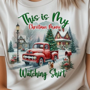 Może przedstawiać: Biała koszulka z czerwonym pickupem, choinką i domem w śnieżnym krajobrazie. Tekst na koszulce brzmi "This is My Christmas Movie Watching Shirt."