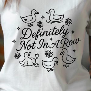 May include: White t-shirt with a black graphic design featuring five ducks, flowers, and the text "Definitely Not In A Row". The design is centered on the front of the shirt.