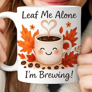 May include: White ceramic mug with the text "Leaf Me Alone I'm Brewing!" featuring a smiling coffee cup, autumn leaves, and coffee beans. The mug is held by a person wearing a white knit hat.