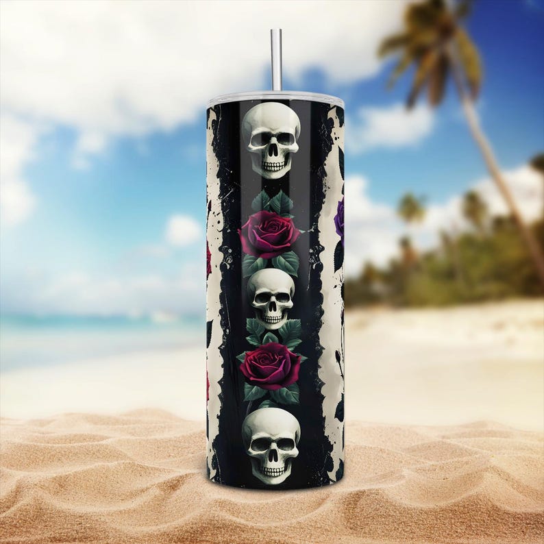 May include: A stainless steel tumbler with a straw, featuring a design of white skulls and red roses. The design is set against a black and white background. The tumbler is on a sandy beach with a blue sky.