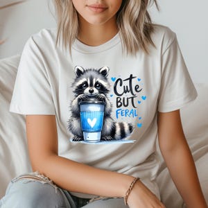 May include: White t-shirt with a cartoon raccoon holding a coffee cup. The text "Cute but Feral" is printed on the shirt.