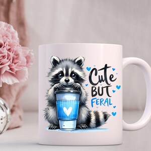 May include: White ceramic mug with a cartoon raccoon holding a coffee cup. The mug has the text "Cute But Feral" in blue lettering.