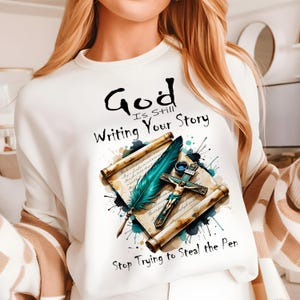 May include: White sweatshirt with a graphic design of a quill pen, an open scroll, and a cross with a figure on it. The text on the sweatshirt reads "God is still writing your story... Stop trying to steal the pen."