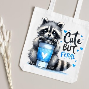 May include: White canvas tote bag with a cartoon raccoon holding a blue and white coffee cup. The text "Cute But Feral" is printed on the bag with blue hearts.