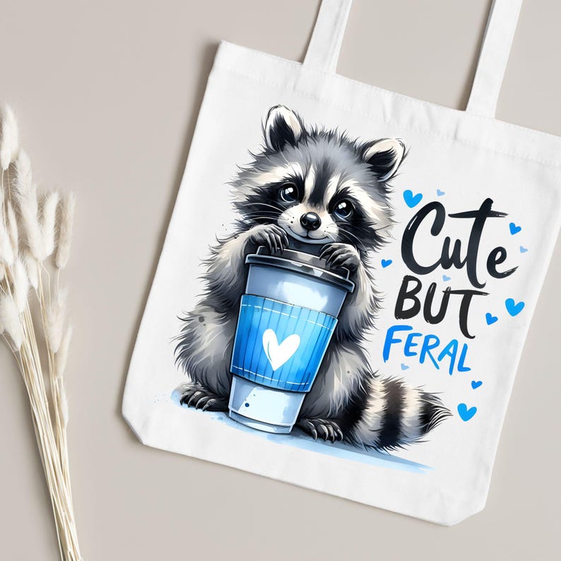 May include: White canvas tote bag with a cartoon raccoon holding a blue and white coffee cup. The text "Cute But Feral" is printed on the bag with blue hearts.