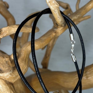 Puede incluir: Un collar de cordón de cuero negro con un cierre plateado. El collar se muestra en una rama de madera.