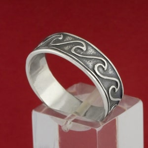 May include: A silver band ring with a wavy design. The ring is on a clear display stand.