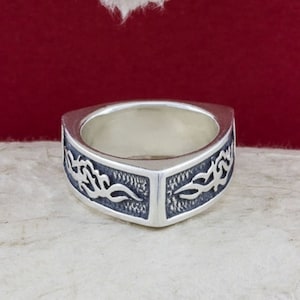 May include: A silver ring with a square band and a detailed design of stylized flames on each side.