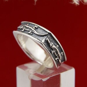 Peut inclure: Une bague en argent avec une bande texturée et un motif de lézards ou de serpents stylisés. La bague est sur un présentoir transparent.