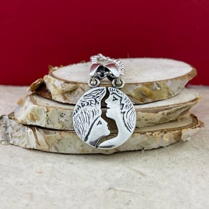 May include: Silver necklace with a circular pendant featuring a detailed design of two faces kissing. The pendant is made of two halves that fit together.