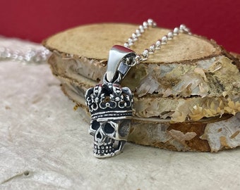 Op de afbeelding: Een zilveren schedel hangerketting met een kroon erop. De hanger zit aan een zilveren ketting en ligt op een stuk hout.