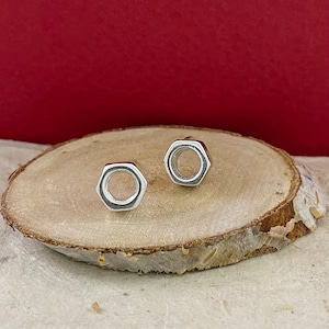 May include: A pair of silver stud earrings shaped like hex nuts. The earrings are on a wooden surface.