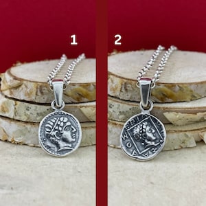 Puede incluir: Dos collares de plata con un colgante de moneda. La moneda de la izquierda presenta un perfil de la cabeza de una persona. La moneda de la derecha presenta un perfil de la cabeza de una persona con texto alrededor del borde.