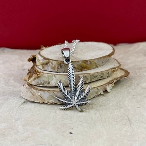 Op de afbeelding: Een zilveren marihuana-blad hangerketting aan een zilveren ketting. De hanger is voorzien van een getextureerd ontwerp en hangt aan een stapel houten rondjes.