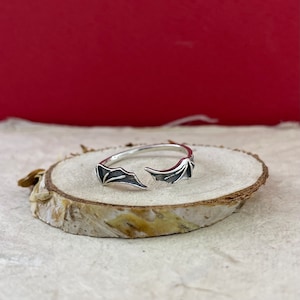 May include: A silver ring with two open wings on the band. The ring is on a light brown wooden surface.