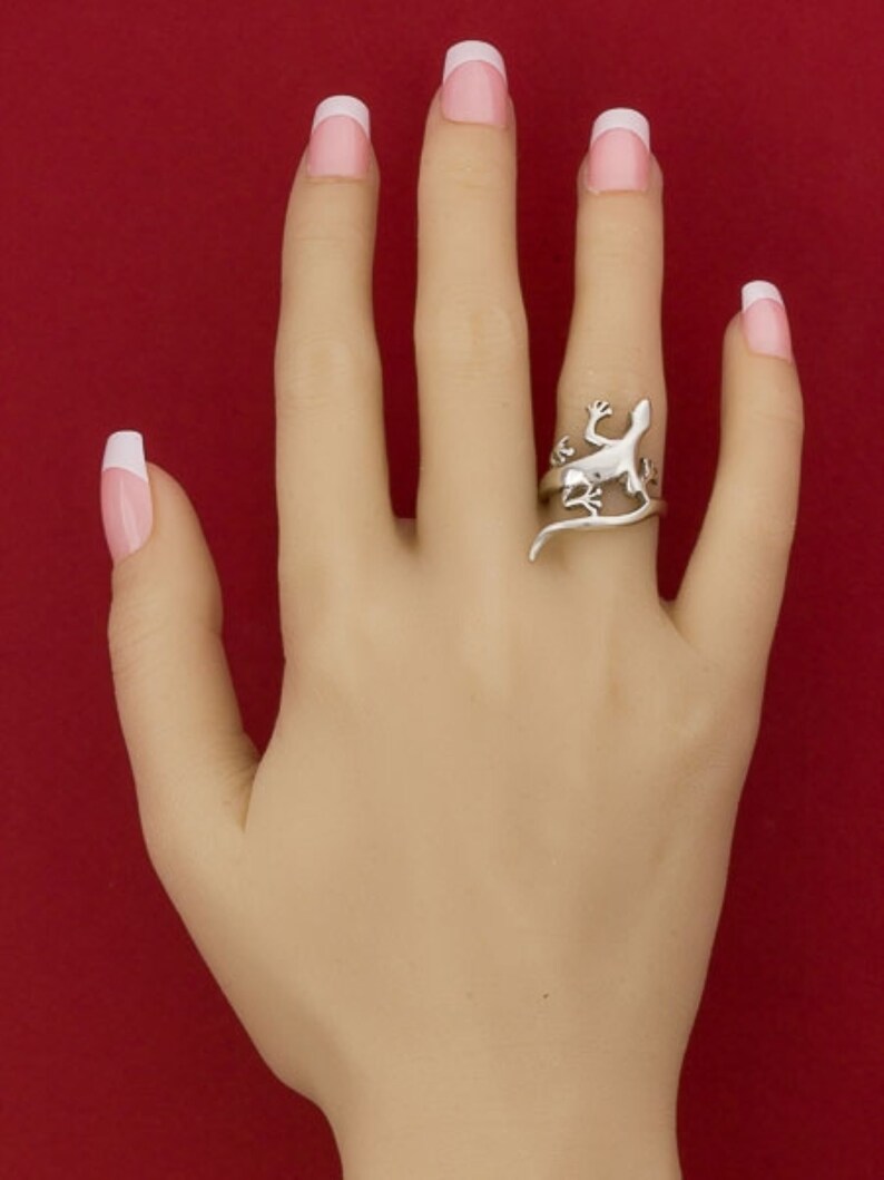 K&ouml;nnte beinhalten: Ein silberner Ring mit einem Eidechsen-Design. Der Ring wird am Mittelfinger einer Hand mit wei&szlig;en French Manik&uuml;re-N&auml;geln getragen.