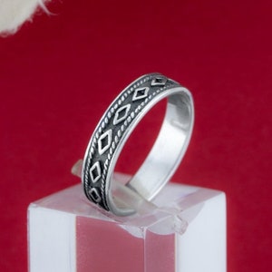 Pode incluir: Um anel de prata com um padrão geométrico em torno da banda. O anel está em um suporte de exibição branco contra um fundo vermelho.