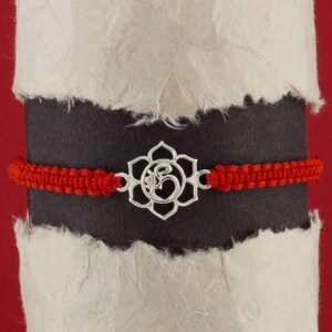 Puede incluir: Pulsera de cuerda roja con un dije de plata con el símbolo Om en forma de flor de loto.