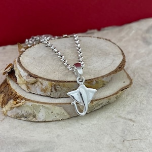 Puede incluir: Un collar plateado con un colgante en forma de manta raya. El colgante presenta un diseño detallado y cuelga de una fina cadena. El collar se exhibe sobre una superficie de madera.