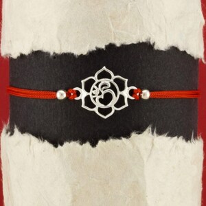 Puede incluir: Un brazalete de cuerda roja con un dije de plata que representa una flor de loto y el símbolo Om.