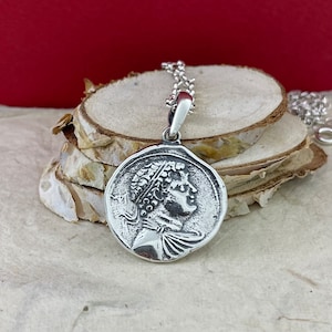 Puede incluir: Collar con colgante de plata que presenta un perfil detallado de una persona con una corona de laurel en la cabeza. El colgante es redondo y tiene una superficie texturizada.