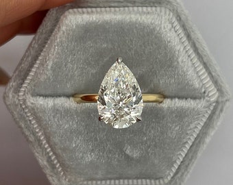 May include: A pear-shaped diamond ring with a gold band, set in a gray velvet hexagonal ring box. The diamond is large and clear, reflecting light. The ring is displayed against a white background.