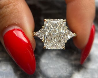 Könnte beinhalten: Ein goldener Ring mit einem Diamanten im Radiant-Schliff. Der Ring hat einen großen rechteckigen Mittelstein, der von zwei kleineren dreieckigen Diamanten flankiert wird. Die Person, die den Ring hält, hat roten Nagellack.