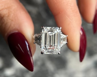 May include: A silver ring with a large, clear emerald-cut diamond and two smaller, triangular diamonds on either side.