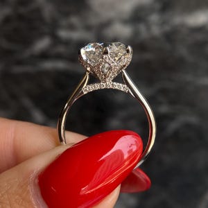 May include: A silver engagement ring with a large, round-cut diamond center stone. The diamond is surrounded by smaller accent stones, and the band is also adorned with small diamonds. The ring is held up against a dark background.