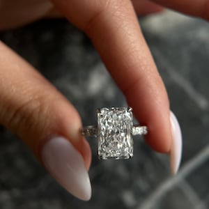 May include: A silver ring with a large, rectangular, clear diamond. The diamond is set in a prong setting and the band is adorned with smaller diamonds.