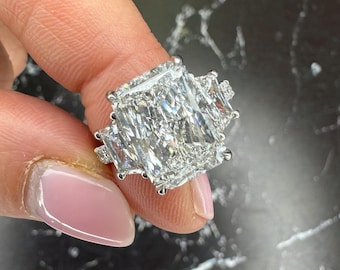 Può includere: Un anello di diamanti con una grande pietra centrale rettangolare e due pietre laterali più piccole. L'anello è incastonato in una fascia d'argento, che mette in risalto la brillantezza dei diamanti. L'anello è mostrato su una superficie di marmo bianco e nero.