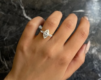 Könnte beinhalten: Ein goldener Verlobungsring mit einem Diamanten im Marquiseschliff. Der Ring hat einen zentralen Diamanten und kleinere Steine auf beiden Seiten. Der Ring wird auf einer Hand mit gepflegten Nägeln präsentiert.