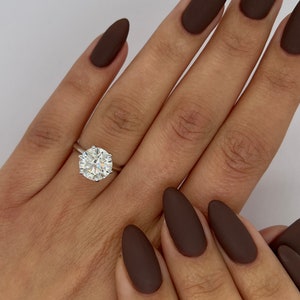 May include: A hand wearing a large, round-cut diamond ring with a silver band. The nails are painted a matte dark brown color. The ring is the focal point, reflecting light.