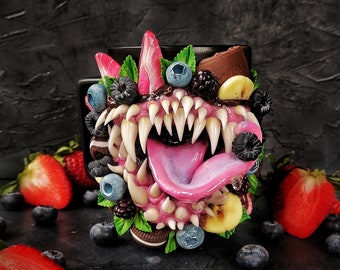 Puede incluir: Una taza de cerámica negra decorada con una cara de monstruo hecha de arcilla polimérica. La boca del monstruo está abierta, revelando dientes afilados y una lengua rosa. La taza está decorada con varias frutas, incluyendo fresas, arándanos y plátanos.