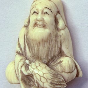 May include: An off-white carved figurine depicting a seated elderly figure with a long beard and a cap, holding a small turtle in their lap. The sculpture features intricate etched details and dark shading within the grooves of the material.