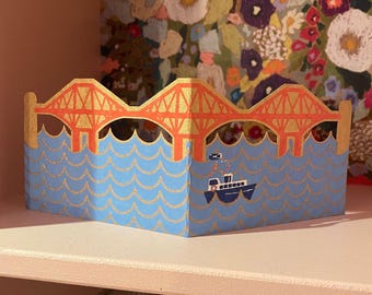 May include: A decorative paper bridge with an orange and gold design, set against a blue water background. A small boat is depicted on the water. The bridge is on a white shelf.