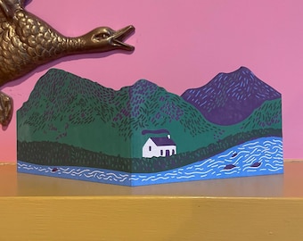 May include: A painted wooden landscape scene featuring a white cottage with a purple roof, green hills, a blue river, and purple mountains. The artwork is displayed on a yellow surface with a bronze duck sculpture in the background.