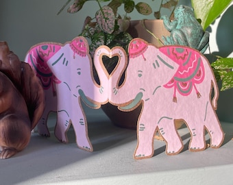 May include: Two pink elephant cutouts with gold glitter accents and decorative patterns. The elephants' trunks form a heart shape. The elephants are decorated with pink, green, and gold patterns. The background includes plants and a frog statue.