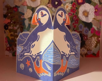 May include: A blue, white, and orange papercraft featuring two puffins. The birds have orange beaks and feet, with blue and white bodies. The base of the papercraft has a blue background with silver wave and star details. A floral painting is in the background.