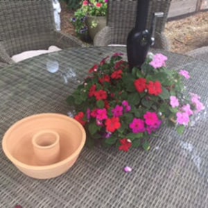 May include: A terracotta planter with a hole in the centre, surrounded by a cluster of pink, red, and white flowers in a glass vase. The planter and vase are on a glass table with a wicker chair in the background.