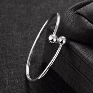 May include: A silver-colored bangle bracelet with two small, round, polished spheres at the opening. The bracelet has a simple, elegant design and is set against a dark background.